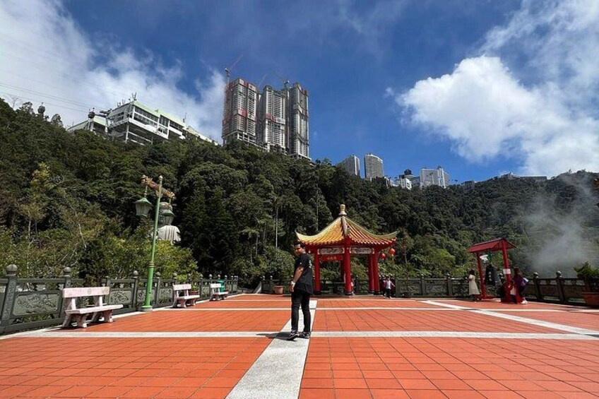 A person standing on a red-tiled walkway with a traditional red pavilion in the background, surrounded by greenery and urban buildings. 4