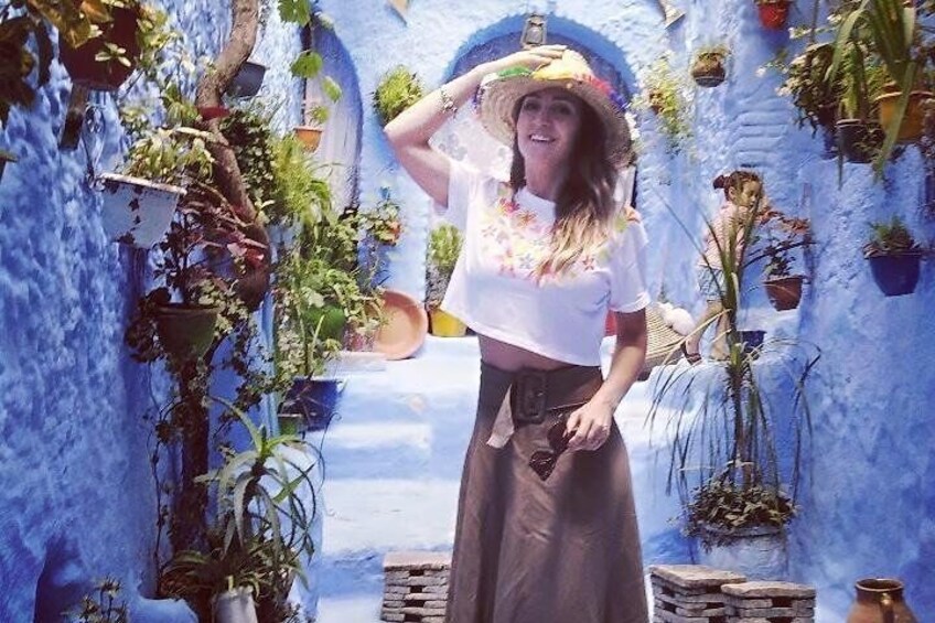 A person in a white top and brown skirt standing in a narrow alleyway with blue walls and potted plants. 1