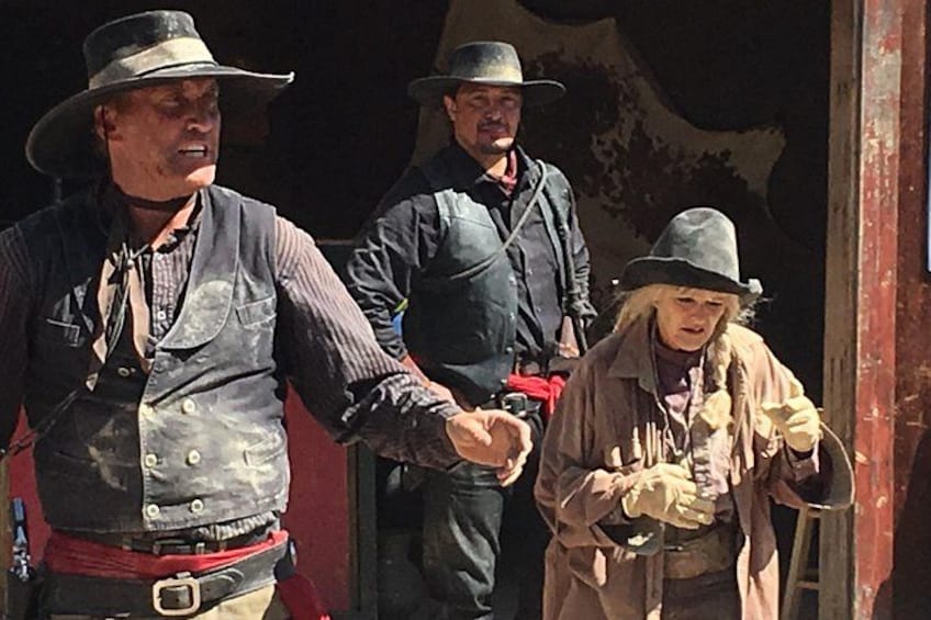 Drie mensen in cowboykleding, waarvan er één een pistool vasthoudt, staan voor een rode deur. 3