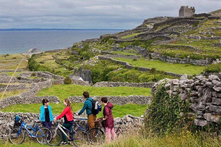A group of people on bicycles, a stone wall, and ruins on a hillside with a view of the sea. 1