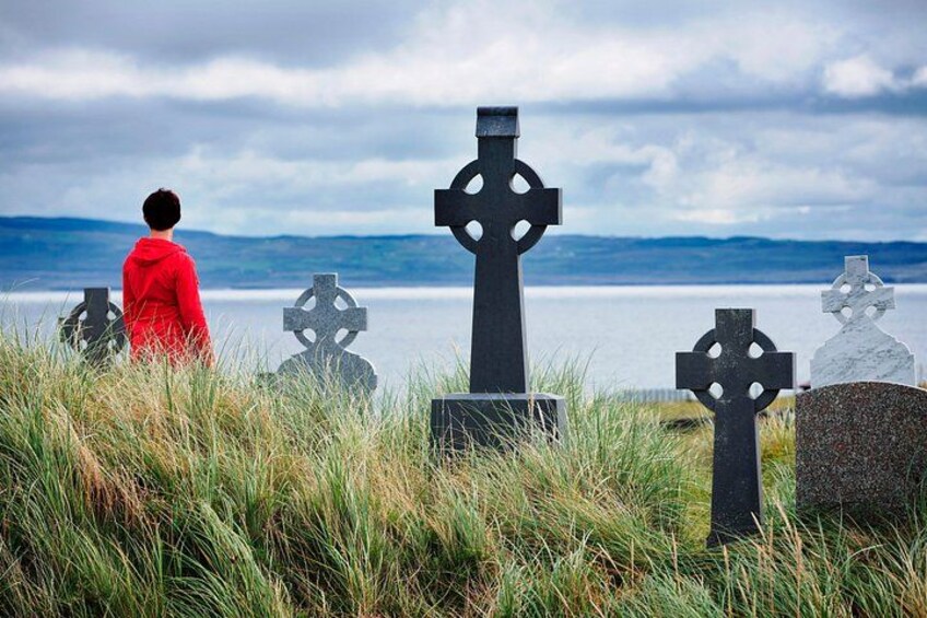 A person in a red jacket standing among gravestones with a lake and mountains in the background. 5