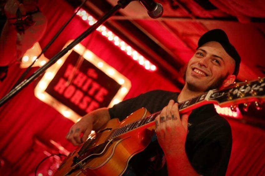 A person playing an acoustic guitar on a stage with a red backdrop and a sign that reads 'WHITE HORSE.' 1
