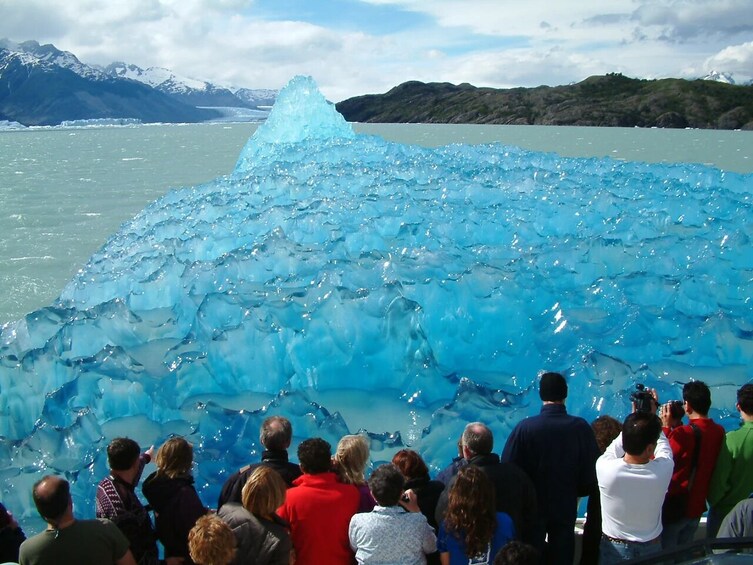 A group of people observing a large, translucent ice formation in a body of water, with mountains in the background. 4