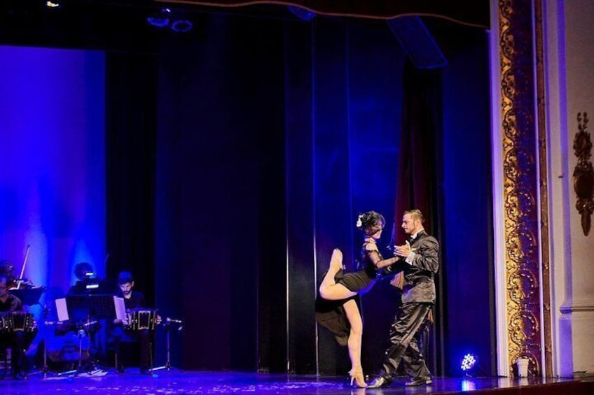 Une scène avec un rideau bleu en arrière-plan, un groupe de musiciens avec leurs instruments, et deux personnes qui exécutent une danse. 3