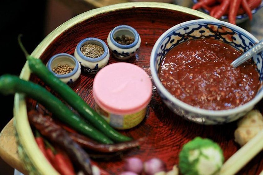 A wooden tray with various spices, a bowl of red sauce, and green chilies. 2