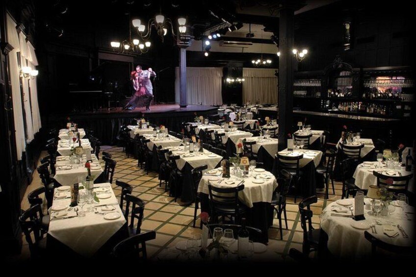 Un restaurante con mesas dispuestas para comer, un escenario con artistas y una zona de bar. 4