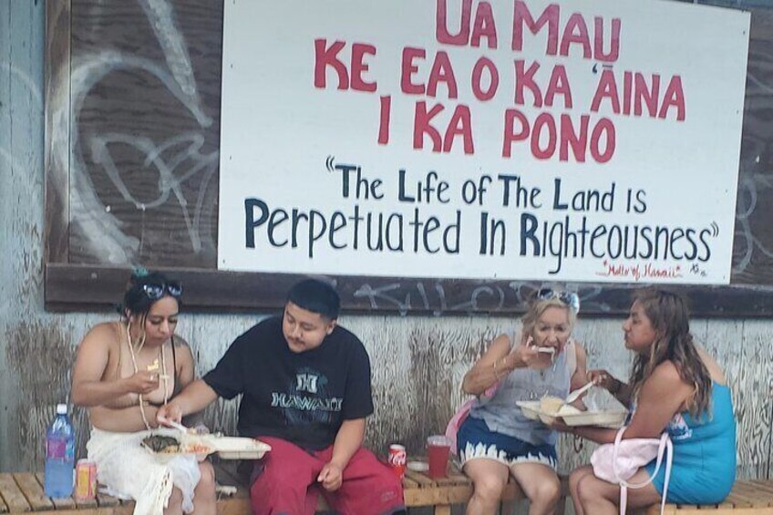 Four people sitting on a bench eating food. A sign with a quote is visible in the background. 1