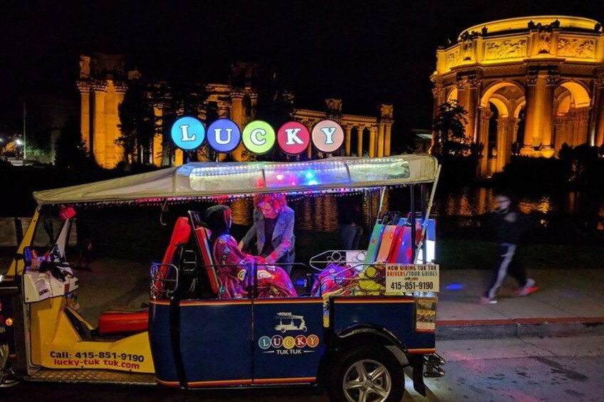 A brightly lit food cart with the word 'LUCKY' on it, parked on a street with a historic building in the background. 1