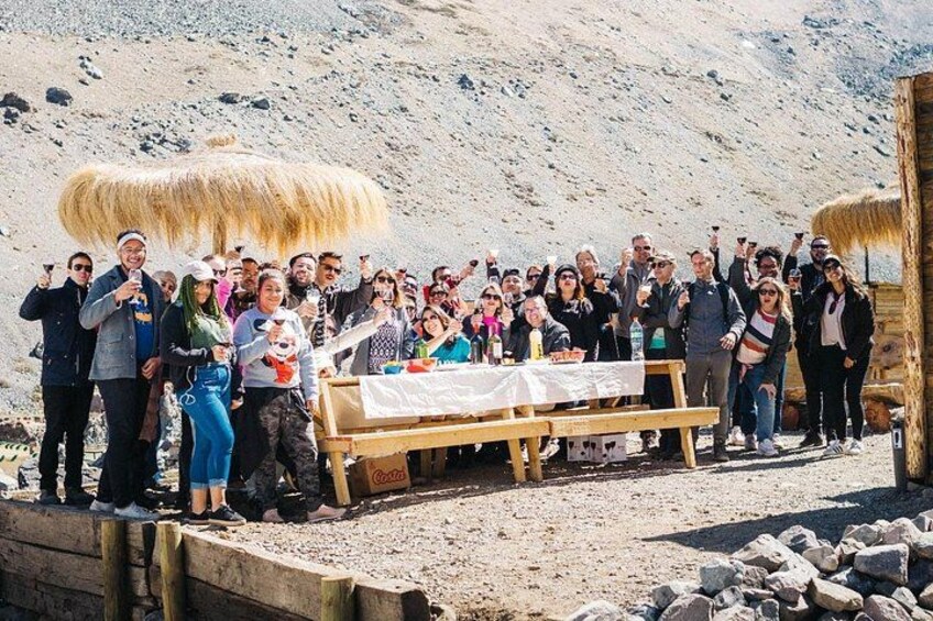 A group of people gathered around a wooden table with a white cloth, standing on a rocky terrain with a mountainous backdrop. 1
