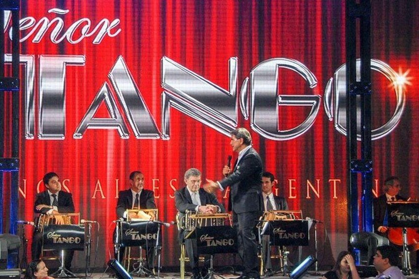Een podium met een rode gordijnen achtergrond en de tekst 'TANGO' in grote letters. Er staan muziekinstrumenten en een spreekgestalte met een persoon. 3