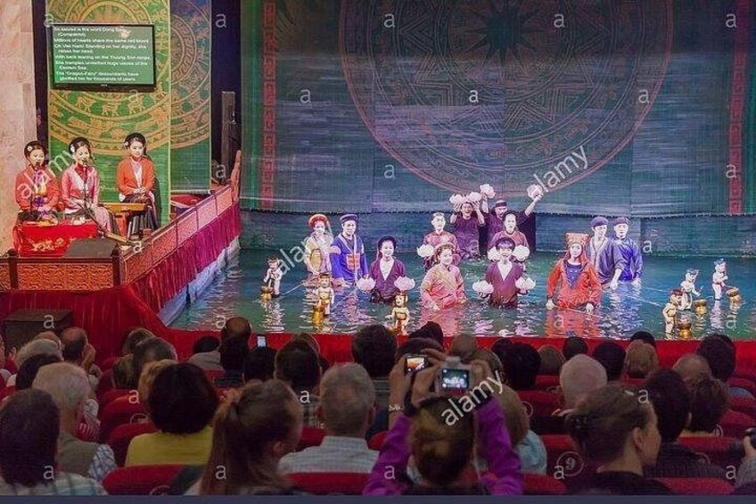Une représentation théâtrale avec des acteurs vêtus de costumes traditionnels, un public et un grand écran en arrière-plan. 5