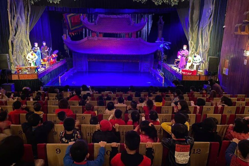 A theater with an audience seated in rows, a stage with performers, and traditional Chinese architectural elements. 2