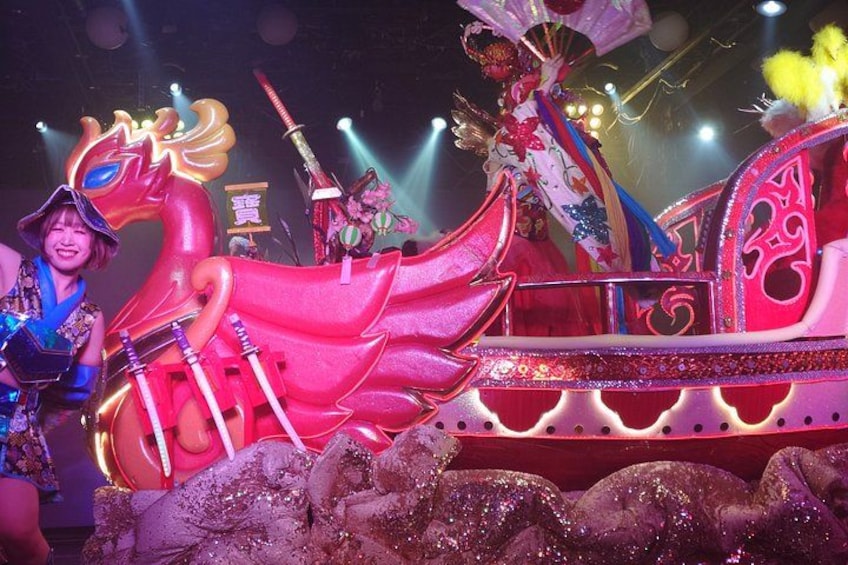 A brightly lit stage with a large, ornate red boat and performers in elaborate costumes. 5