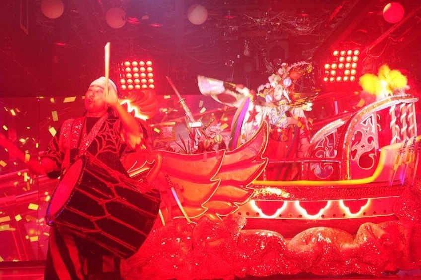 A performer in a red outfit with a large bag, a stage with a red and gold themed backdrop, and a decorative carriage. 4
