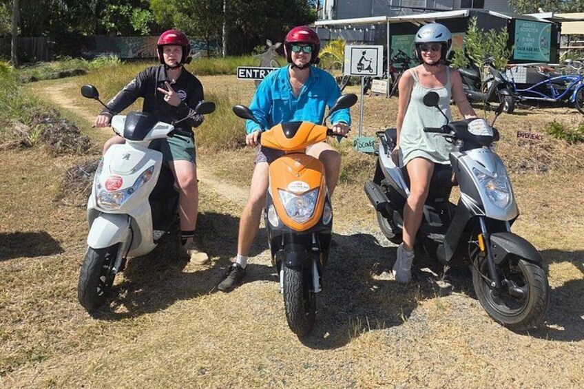 Three people on scooters on a dirt path with a sign in the background. 1