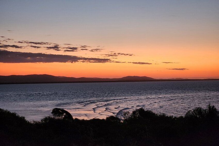 A sunset over a calm sea with waves, distant mountains, and silhouetted vegetation. 5