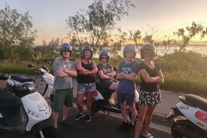 A group of people standing by motorcycles on a road with trees and a sunset in the background. 2