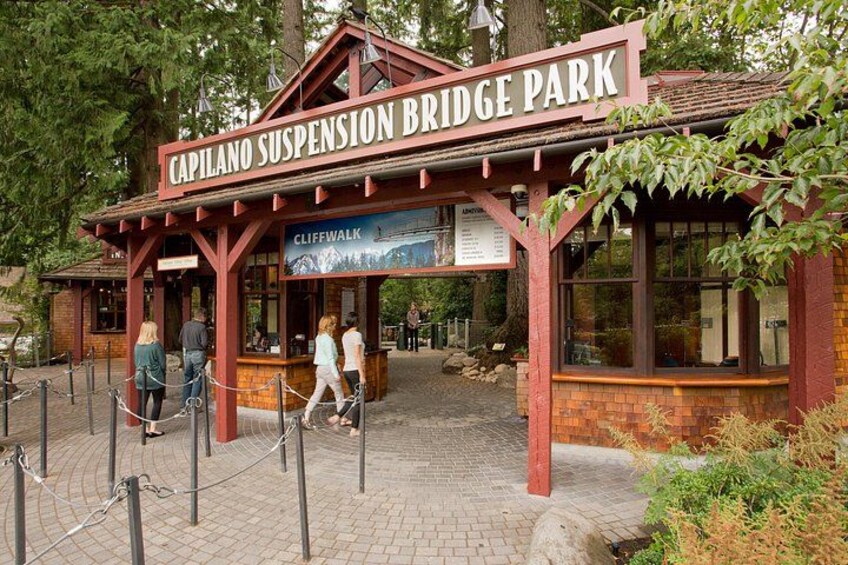 A suspension bridge park with a red wooden structure, a sign reading 'Capilano Suspension Bridge Park,' and a 'Cliffwalk' sign. 1