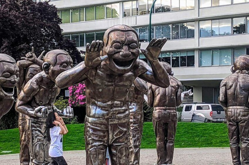 Een aantal bronzen beelden van mensen in verschillende houdingen, met een gebouw en voertuigen op de achtergrond. 5
