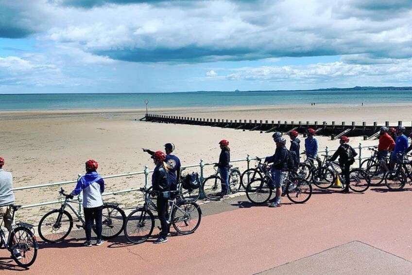 A group of cyclists on a paved path by a beach with a wooden jetty extending into the water. 3