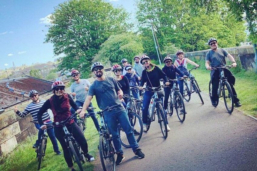 A group of cyclists on a paved path with greenery and a stone wall in the background. 1