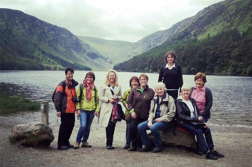 A group of people standing and sitting by a lake with mountains in the background. 2