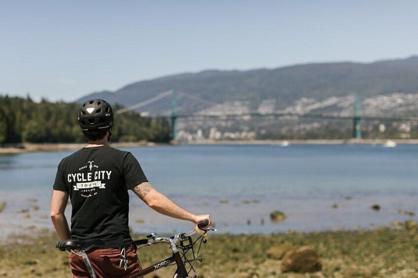 A person on a bicycle by a lake with a bridge in the background. 3