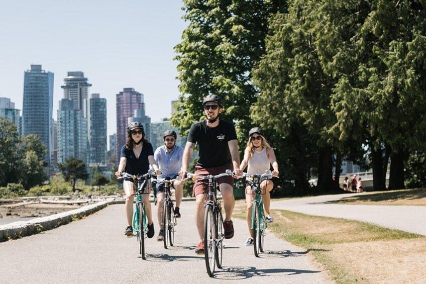 En gruppe mennesker cykler på en belagt sti i en park med træer og en bys skyline i baggrunden. 3