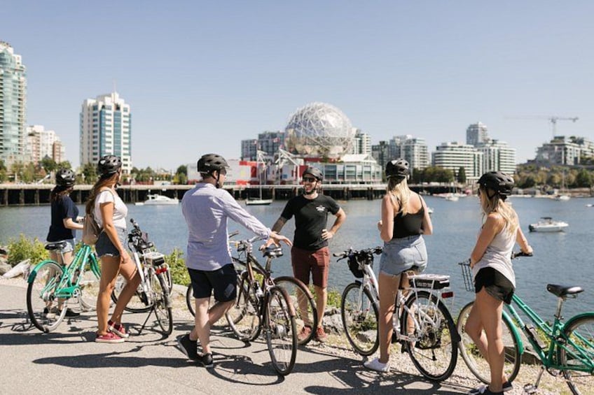 A group of people with bicycles by a waterfront, with buildings and a dome structure in the background. 2