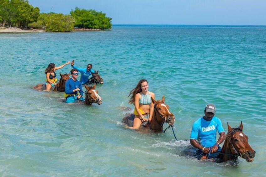 People riding horses in shallow water with a clear view of the ocean and distant land. 1