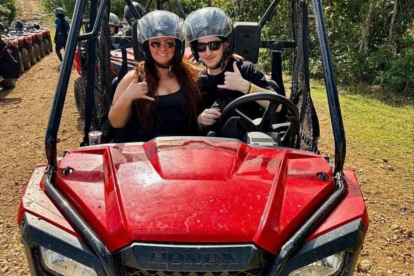 A red ATV with two people wearing helmets and sunglasses, a dirt path, and trees in the background. 3