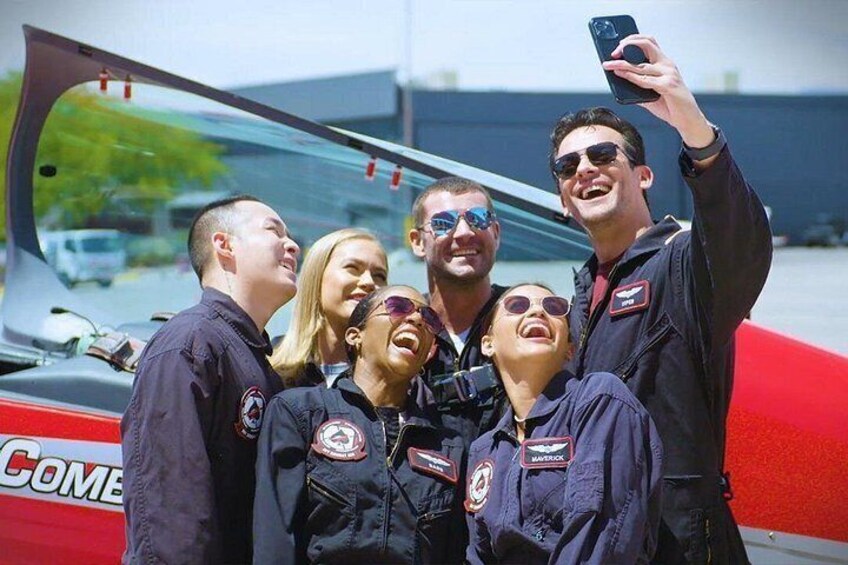 A group of people in flight suits taking a selfie with a red aircraft in the background. 2