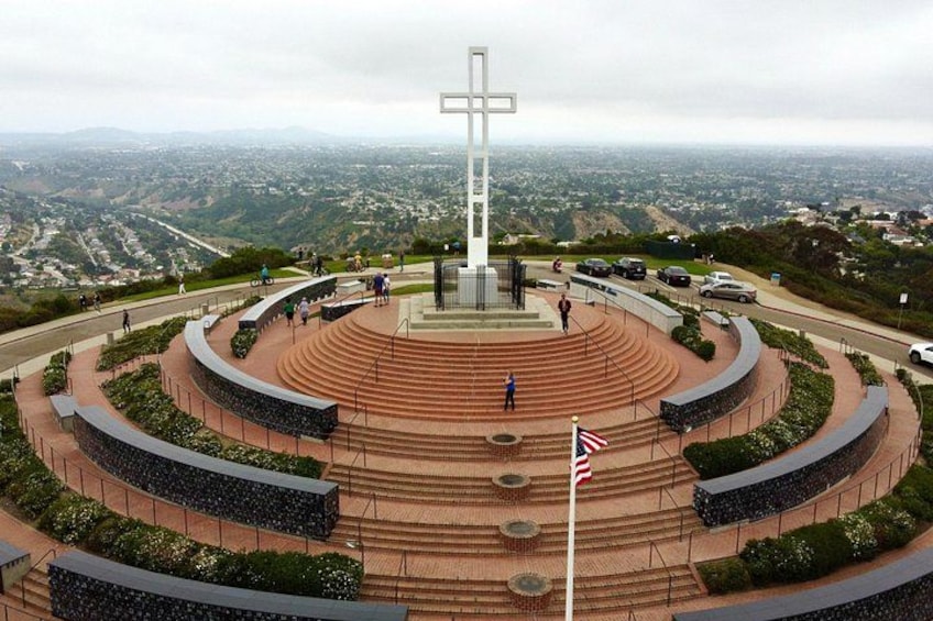 A memorial with a cross, surrounded by steps and greenery, overlooking a cityscape. 1