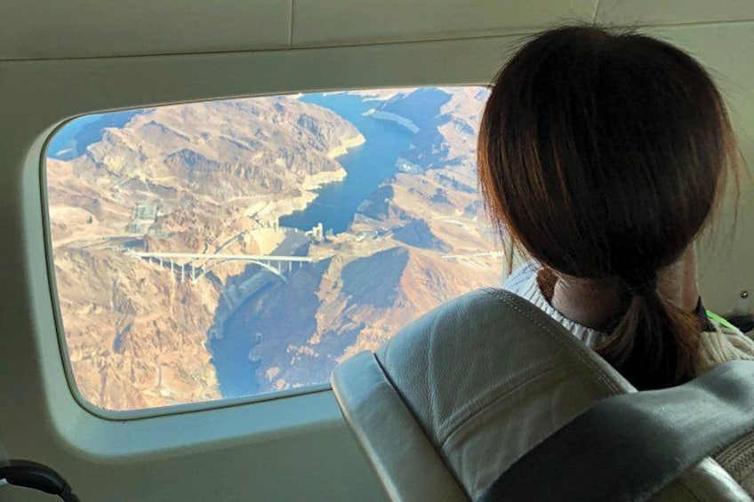 A person seated inside a vehicle, looking out through a window at a bridge over a river, with dry, arid land surrounding the area. 2