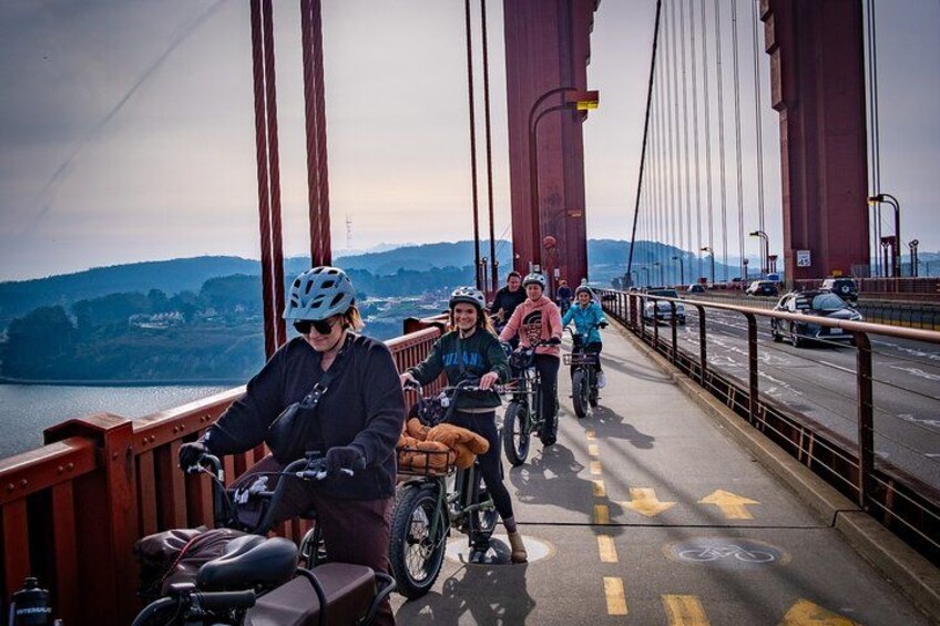 Des personnes à vélo traversent un pont d’où l’on peut admirer un paysage pittoresque, avec une étendue d’eau et des collines en arrière-plan. 2