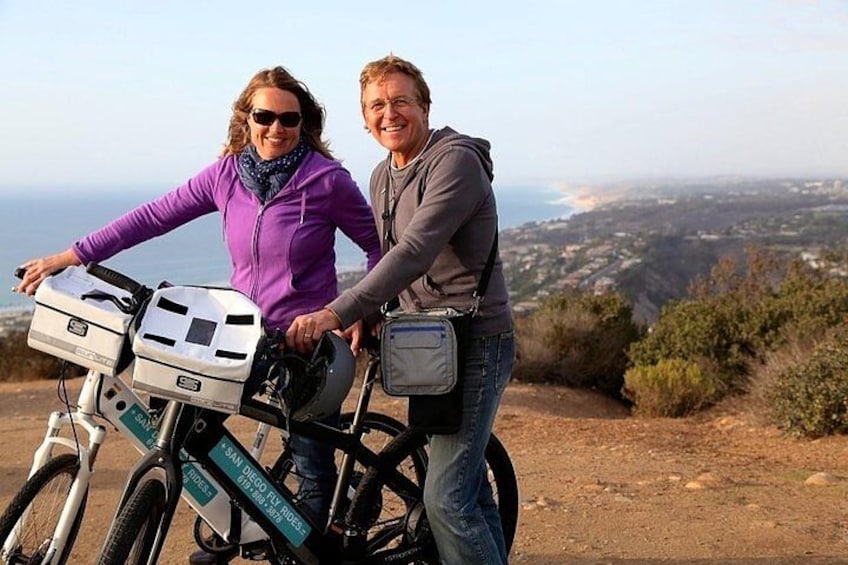 Two people on a bicycle with a map and a bag, overlooking a landscape with hills and a distant view of a city. 3