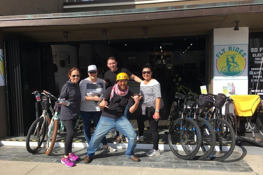 A group of people posing in front of a bicycle shop with bicycles parked outside. 2