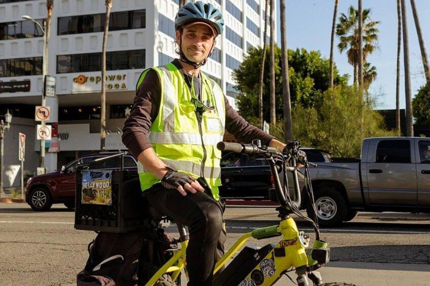 Una persona en un patinete eléctrico amarillo con una caja de entrega, usando un chaleco de seguridad, en un entorno urbano con palmeras y edificios. 2