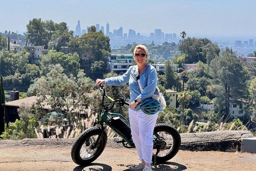 Una persona de pie junto a una bicicleta eléctrica verde, con un paisaje urbano al fondo. 3