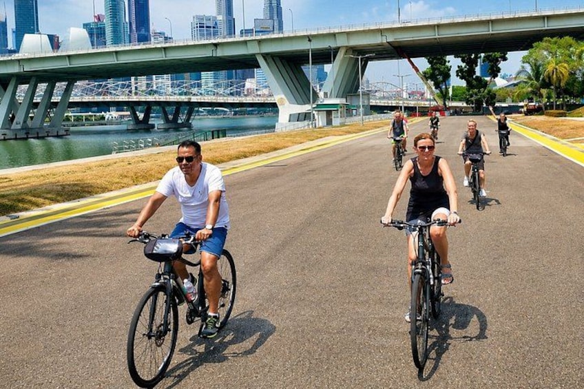A group of cyclists on a paved path with a city skyline and a bridge in the background. 3