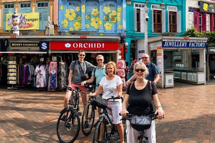A group of people on bicycles in a shopping area with storefronts like 'Money Changer' and 'Orchid!' 5