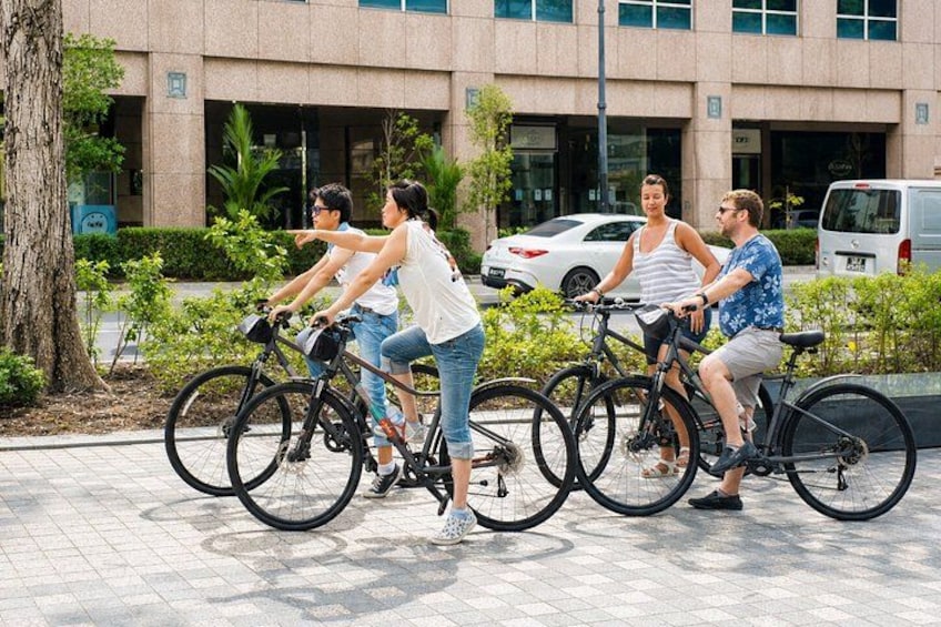 Four people riding bicycles on a paved path with a building and parked cars in the background. 4