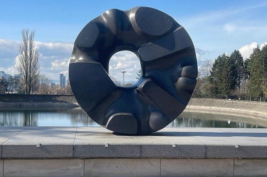 A large, abstract sculpture with a circular design, located near a body of water with a cityscape in the background. 2