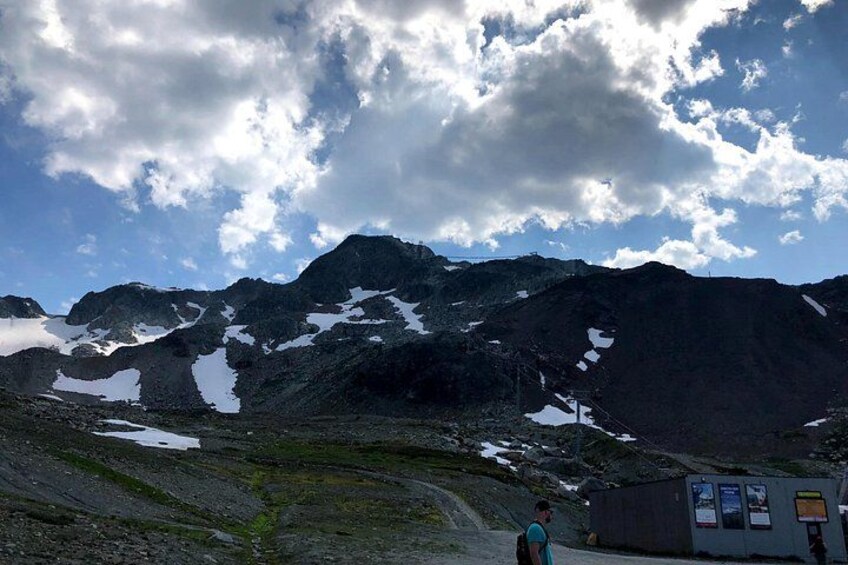 A mountainous landscape with snow patches, a person standing, and a small building with informational posters. 3