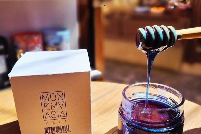 A honey dipper dripping honey into a jar, a cardboard box with 'MON EMVA DELI' text, and a blurred background. 3