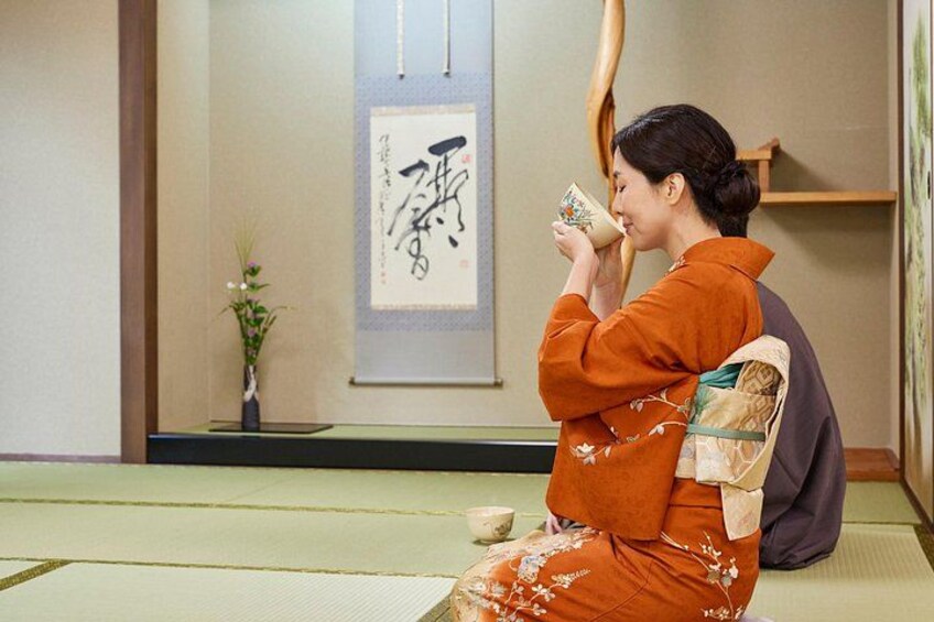 A person in traditional attire sitting on tatami mats in a room with a calligraphy piece on the wall. 2