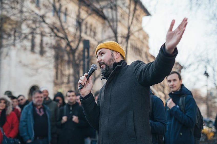 A person speaking into a microphone with an audience in an outdoor setting. 2