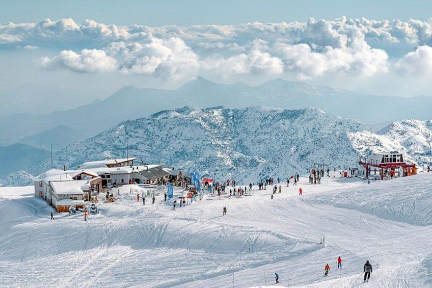 A snow-covered ski slope with skiers, a bus, and buildings in the background. 2