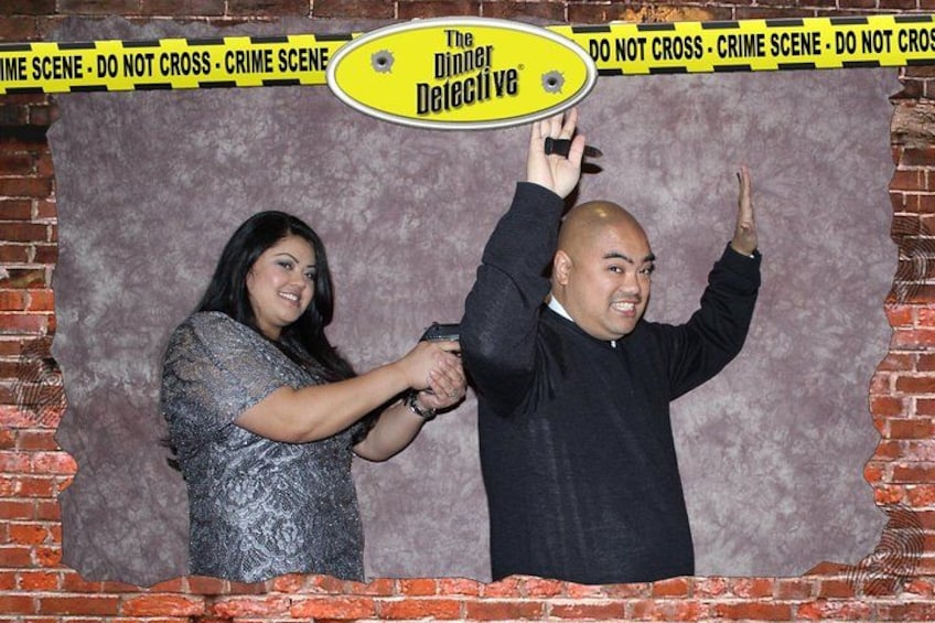 Two people in front of a backdrop with 'The Dinner Detective' text and 'DO NOT CROSS - CRIME SCENE' tape. 2