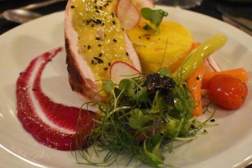 Een bord eten met een stuk vlees, overgoten met gele saus, versierd met kruiden en microgroenten, vergezeld van rode saus en diverse groenten. 1